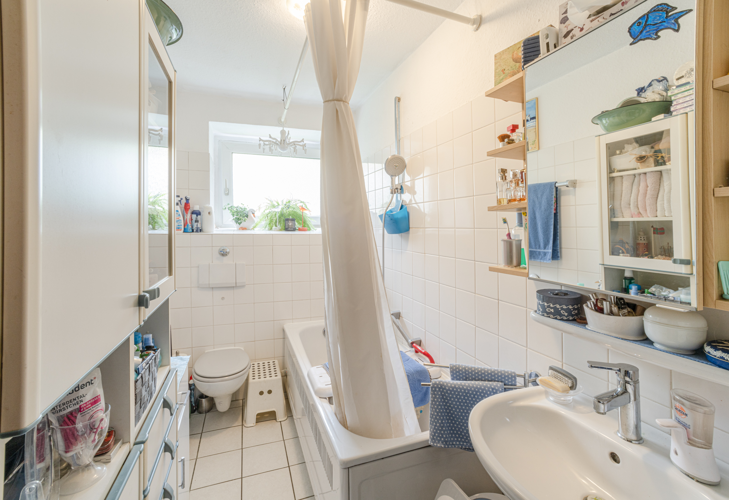 Helles Badezimmer mit Badewanne, Duschvorhang, Waschbecken, WC und Regal an der Wand.