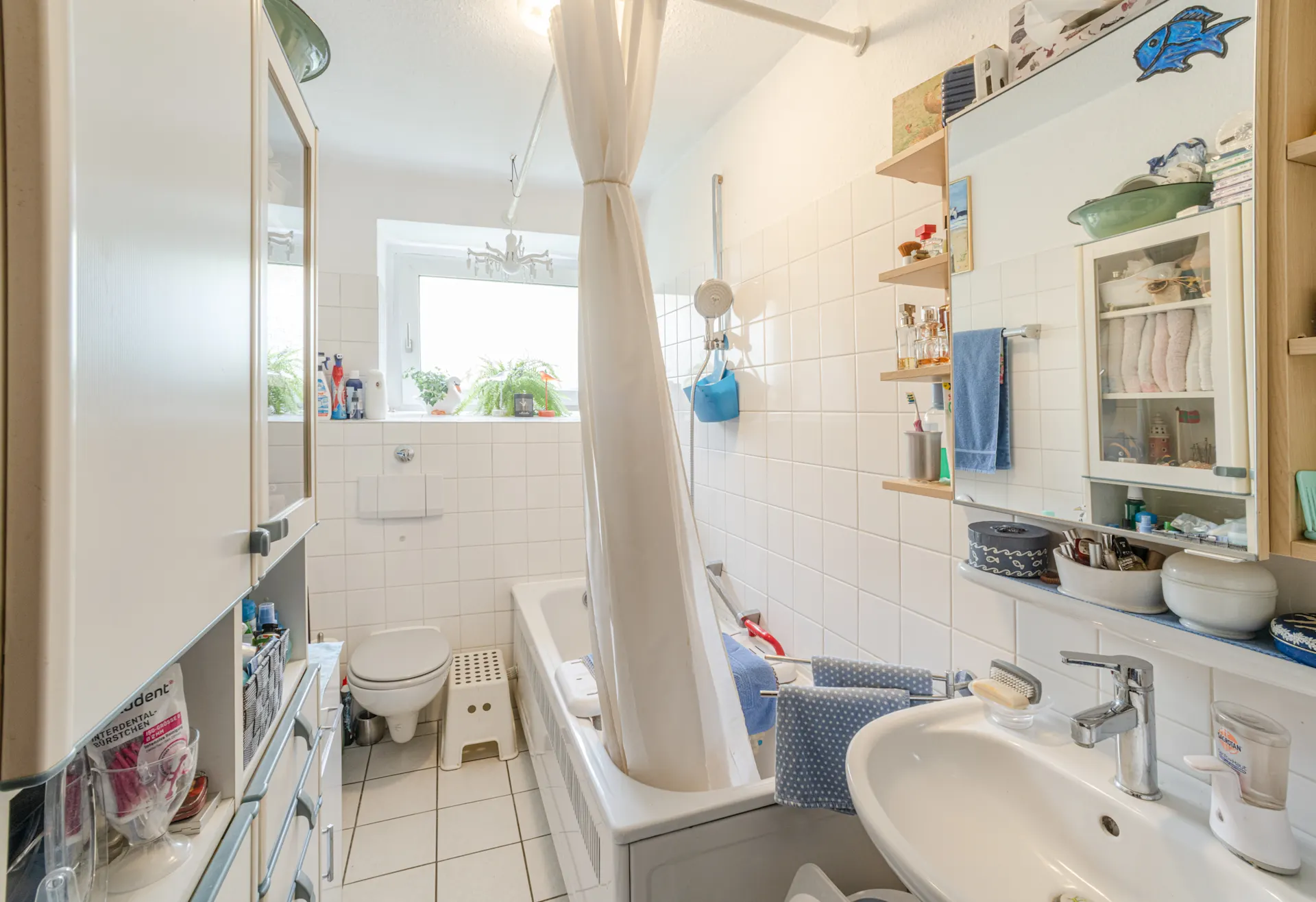 Helles Badezimmer mit Badewanne, Duschvorhang, Waschbecken, WC und Regal an der Wand.