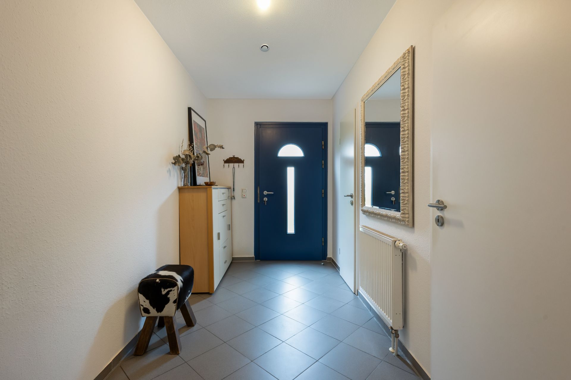 Heller Flur mit blauer Tür, Spiegel, Kommode und Hocker auf gefliestem Boden.