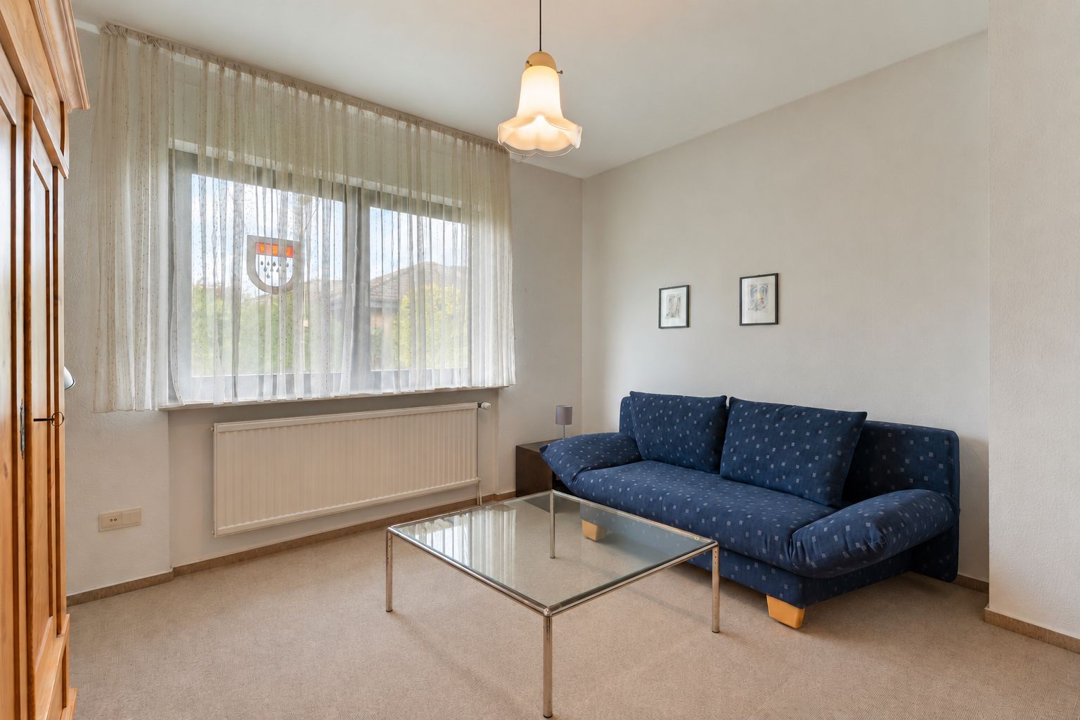 Wohnzimmer mit blauem Sofa, Glastisch und großem Fenster mit Vorhängen.