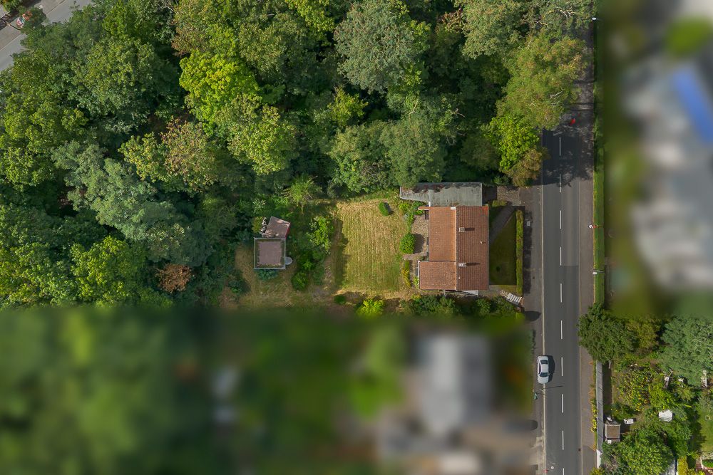 Luftaufnahme eines Wohnhauses mit Garten und angrenzender Straße, umgeben von vielen Bäumen.