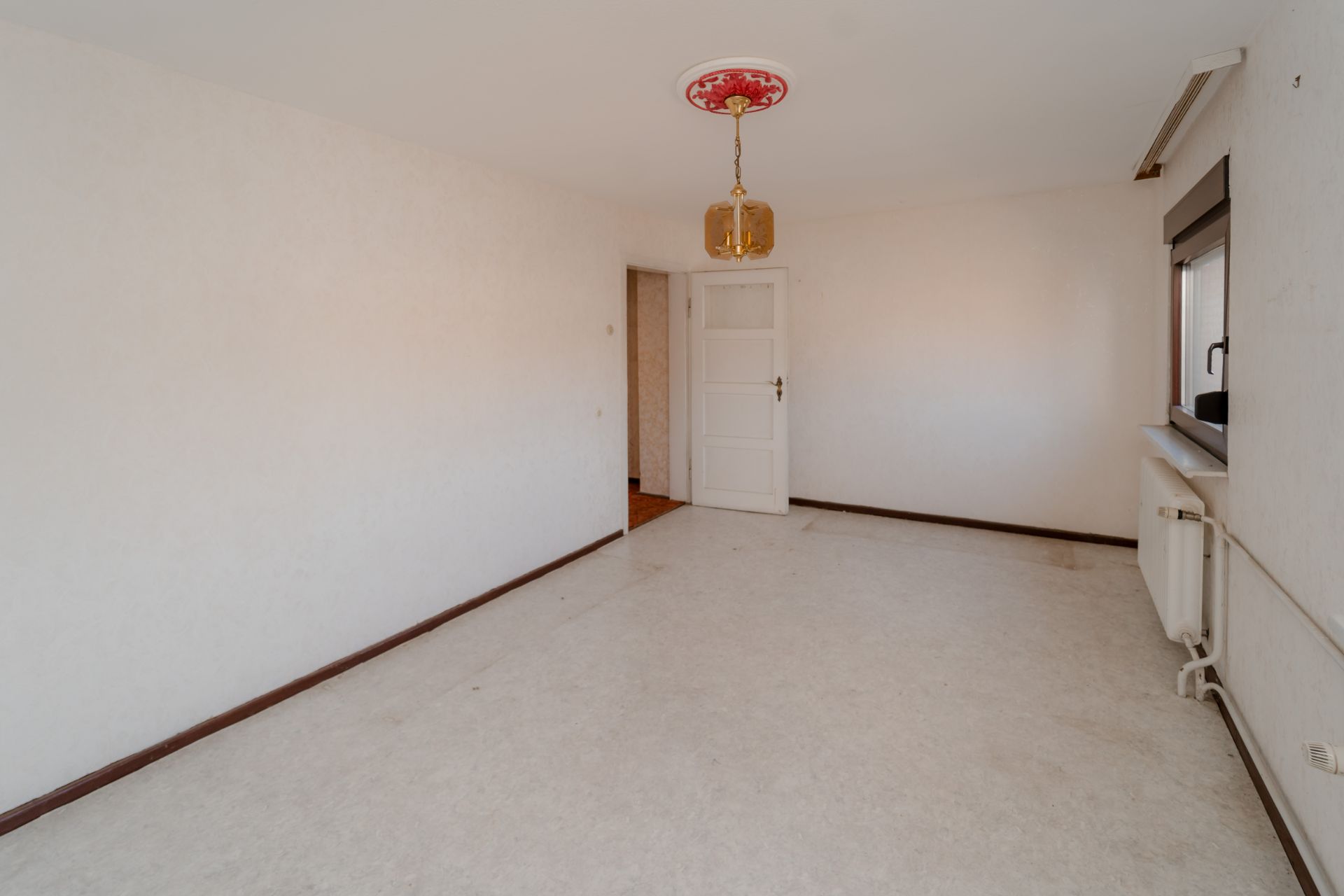 Leeres Zimmer mit weißer Tür, Fenster, Heizung und Lampe an der Decke.