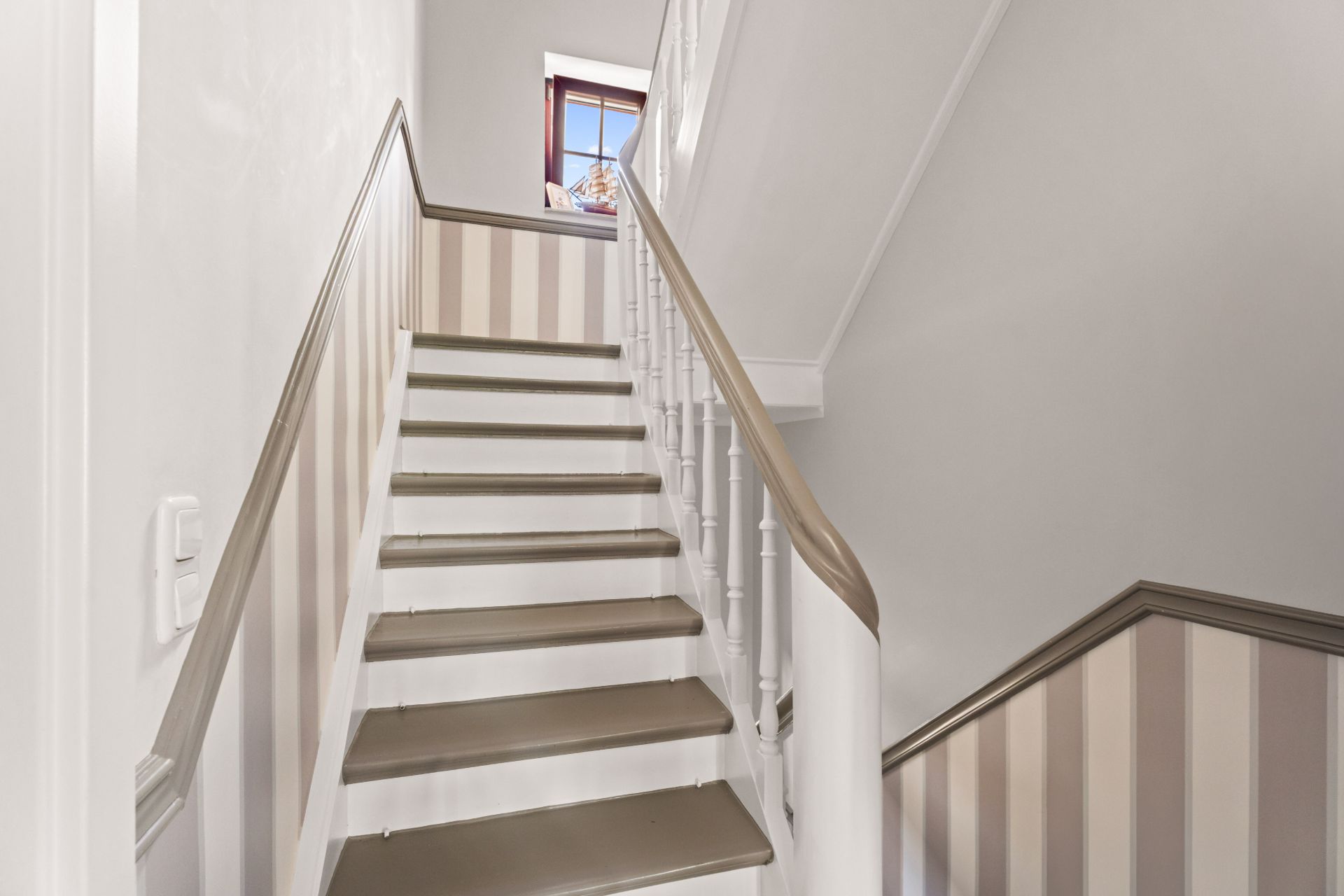 Eine weiß gestrichene Treppe mit Holzgeländer und gestreifter Wandverkleidung, oben ein Fenster.