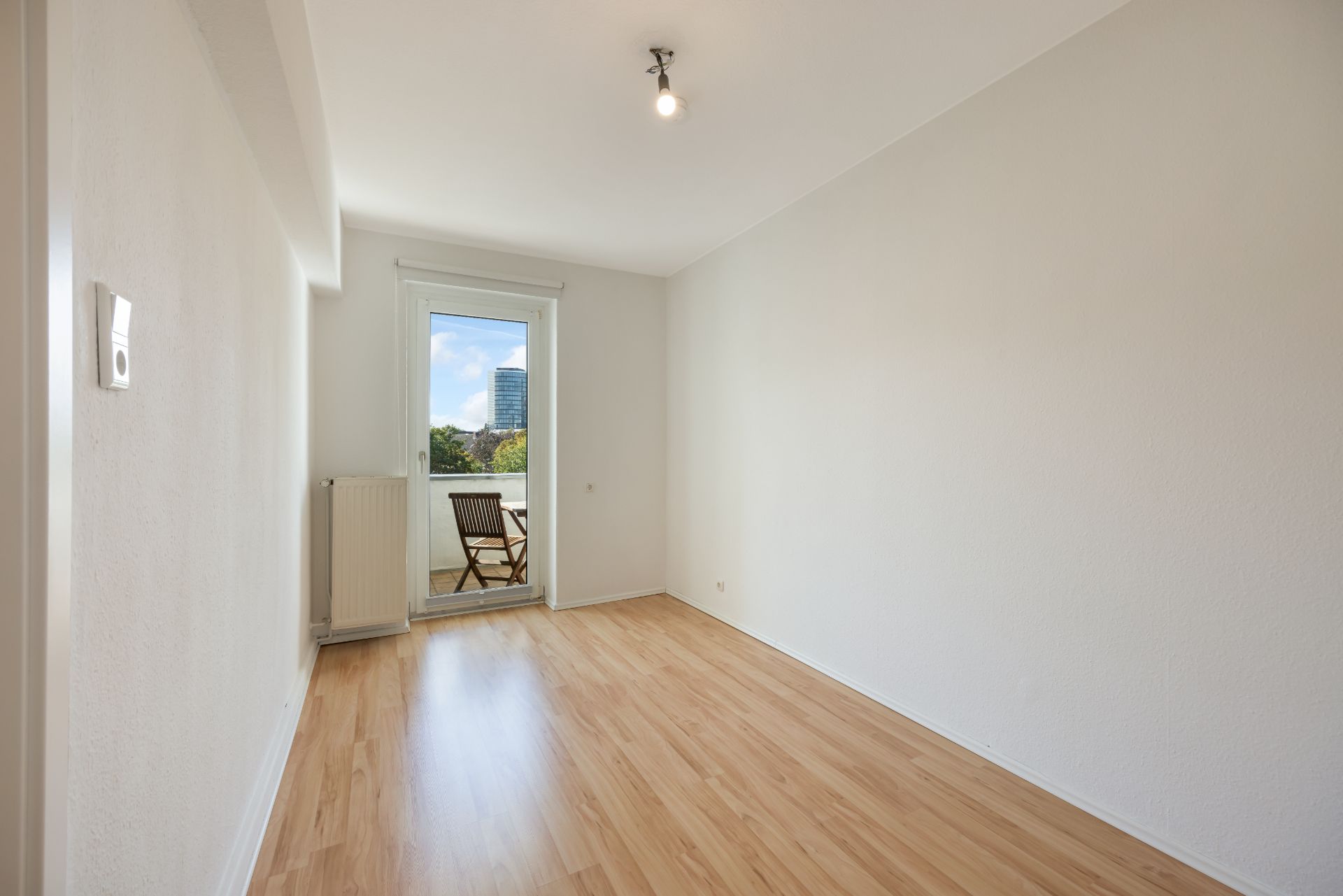 Leeres Zimmer mit Holzfußboden und Zugang zu einem Balkon mit Stuhl und Tisch.