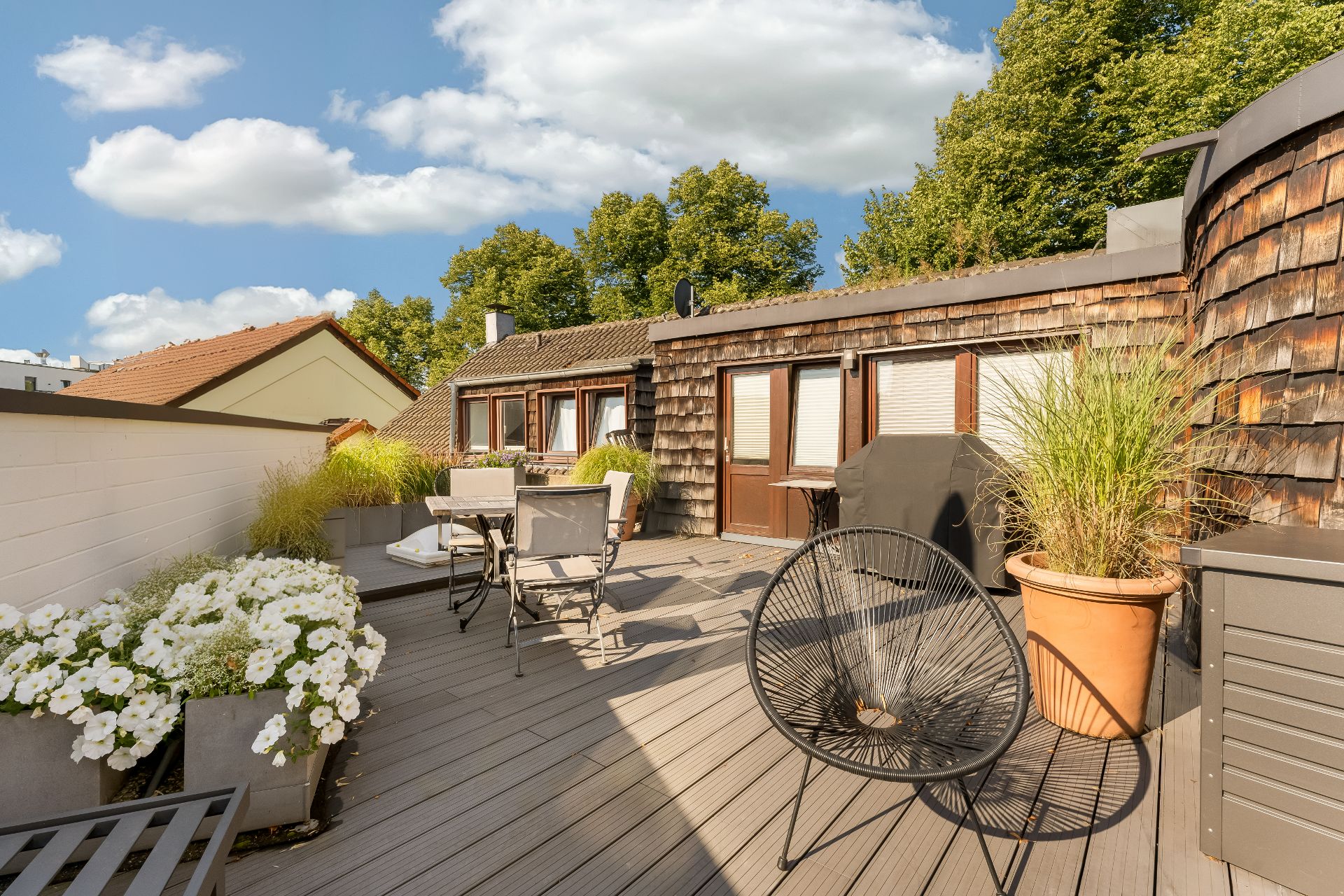 Große Terrasse mit Blumen, Stühlen, Tisch, Grill und Pflanzen, Holzwand und blauer Himmel.