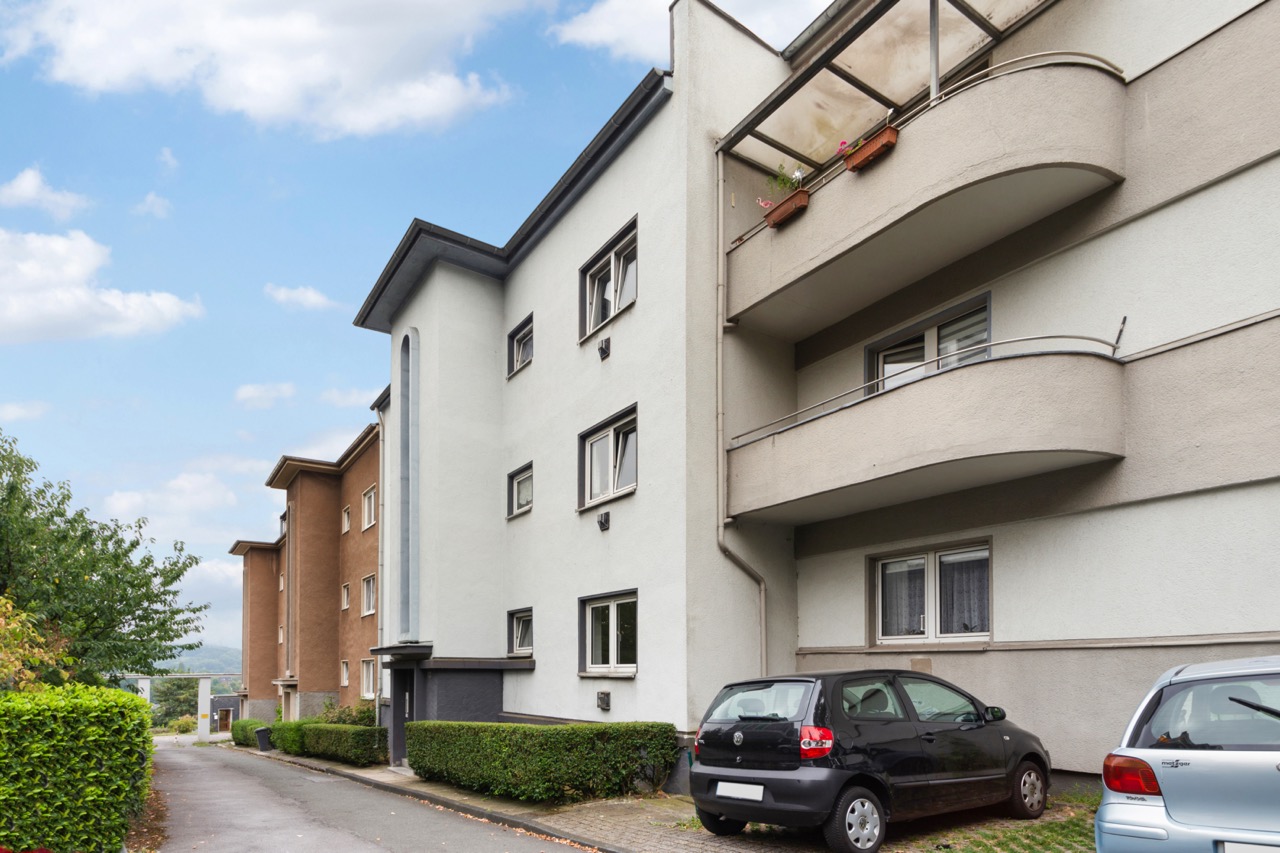 Mehrfamilienhaus mit Balkonen, geparkte Autos davor und Gehweg mit Büschen.