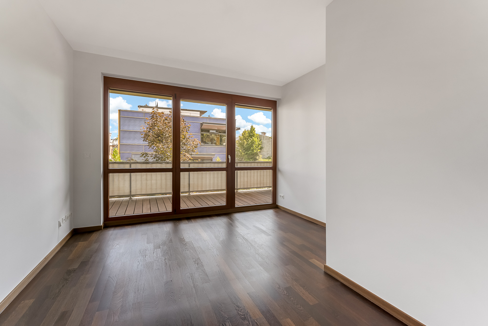Leeres Zimmer mit dunklem Holzboden und großen Fenstern, die auf einen Balkon führen.