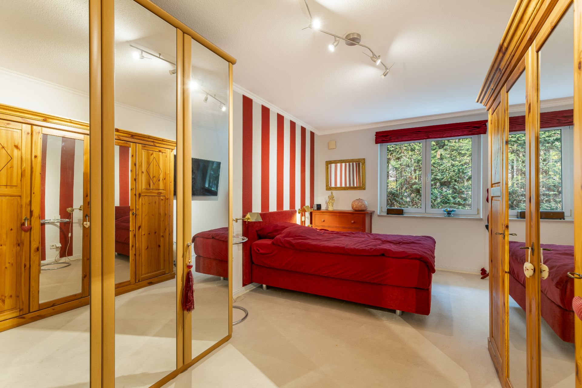 Schlafzimmer mit rotem Bett, Kleiderschränken mit Spiegeln und Fenstern, gestreifte Wand im Hintergrund.