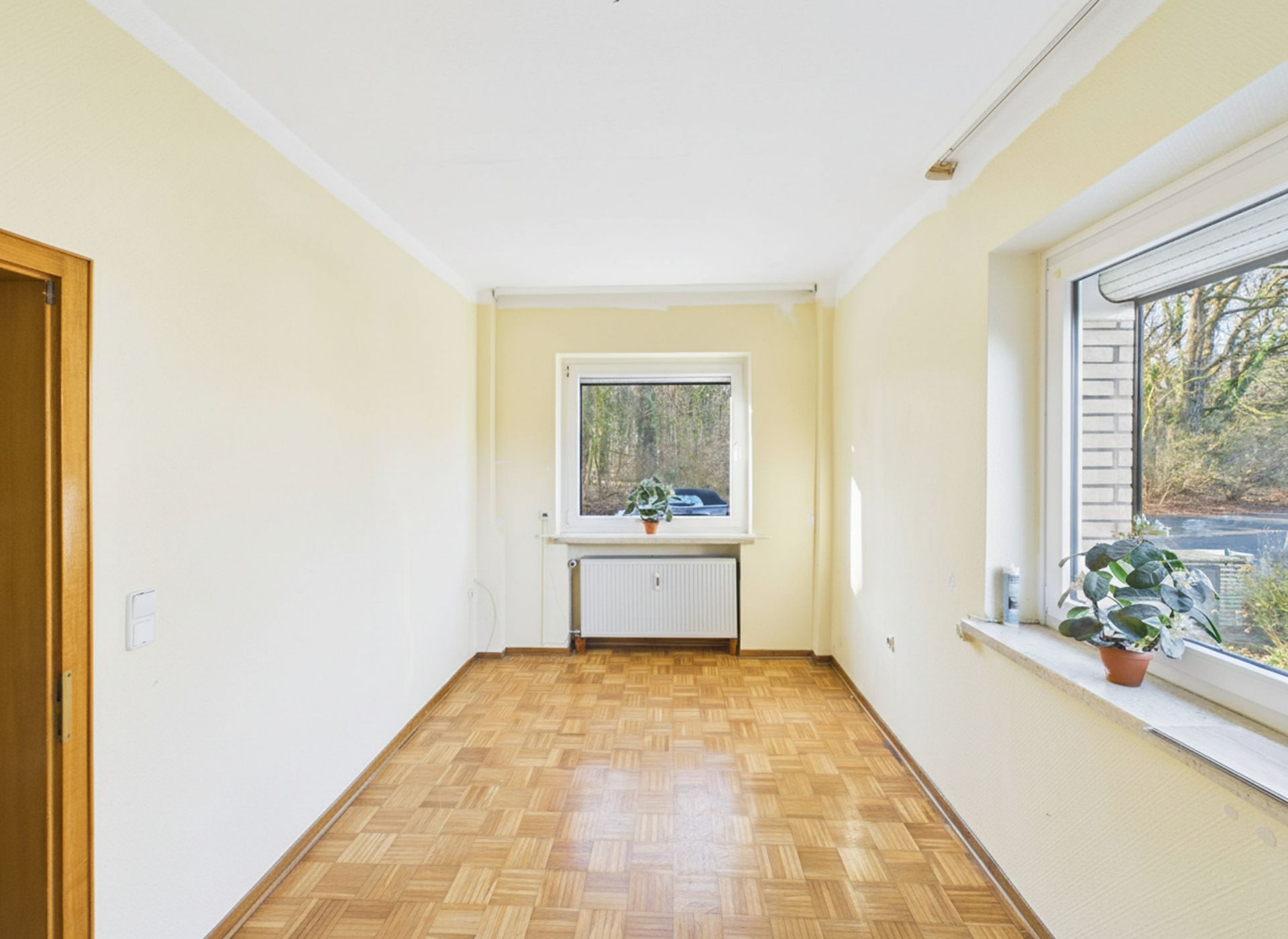 Heller Raum mit Parkettboden, zwei Fenstern und zwei Zimmerpflanzen auf Fensterbänken.