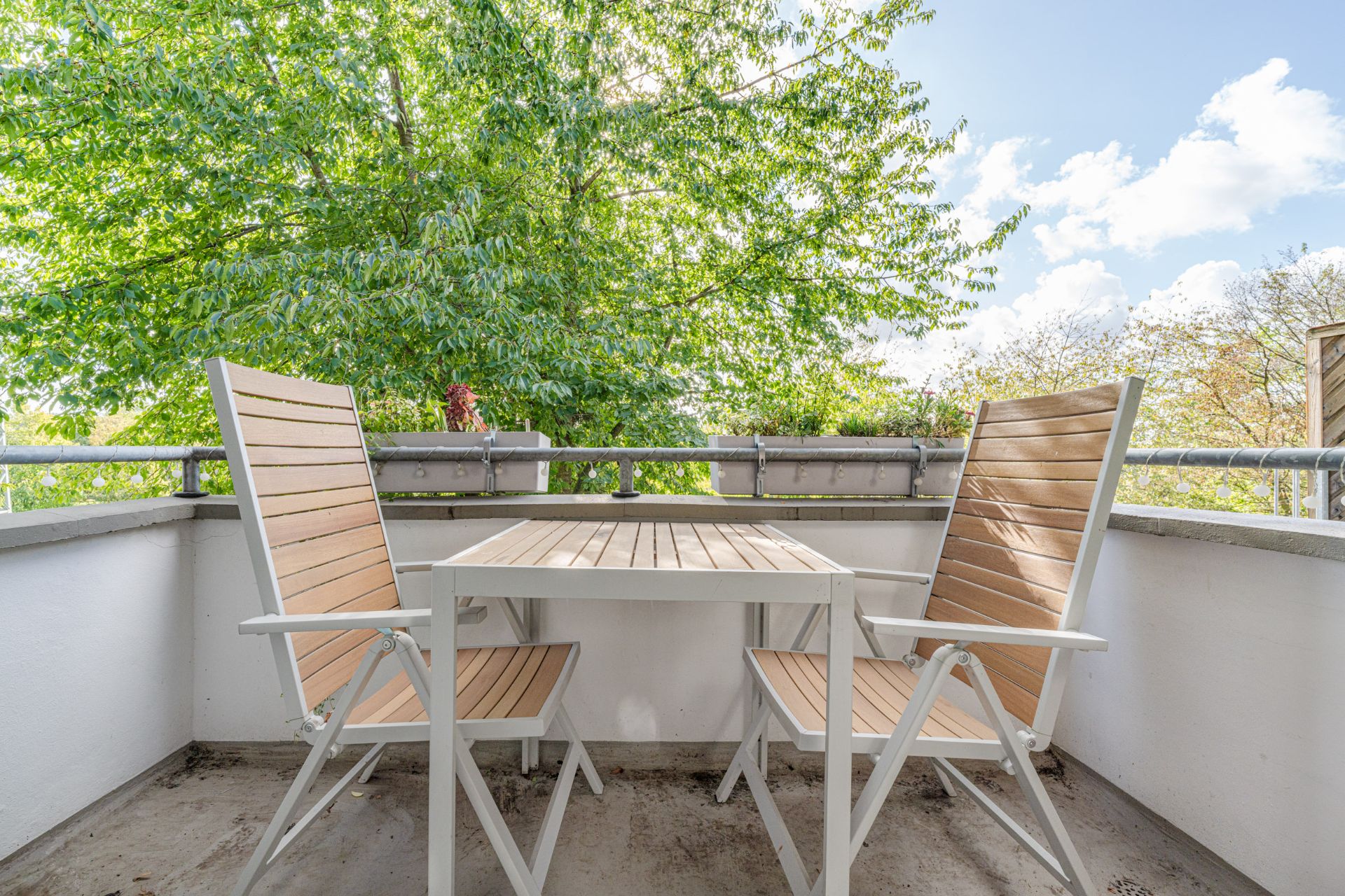 Zwei Holzstühle und ein Holztisch auf einem Balkon mit Blick auf grüne Bäume.