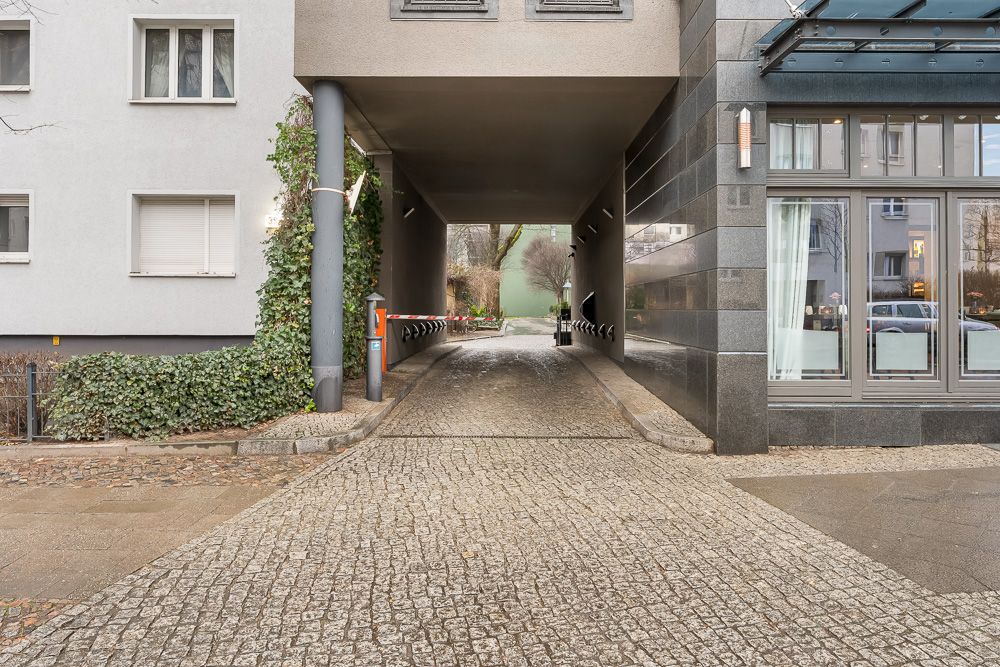 Einfahrt mit Schranke zwischen zwei Wohngebäuden, gepflasterter Weg und einige Pflanzen.