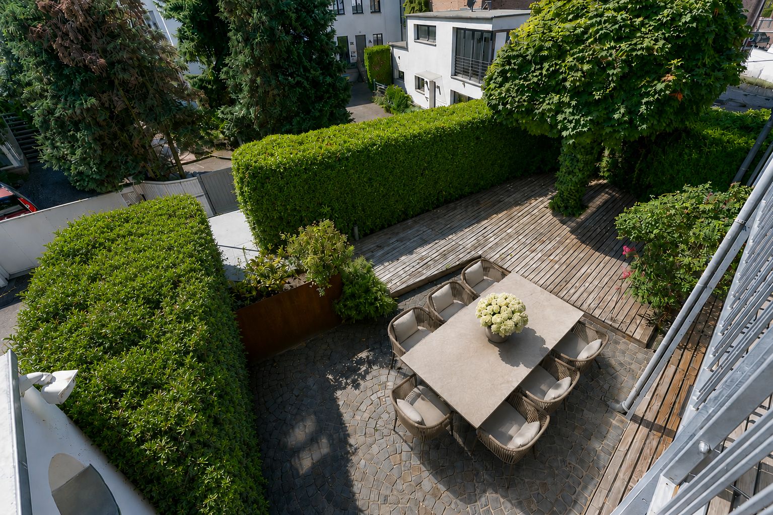 Gepflegter Innenhof mit Steinterrasse, Esstisch und Stühlen, umgeben von grünen Hecken und Bäumen.