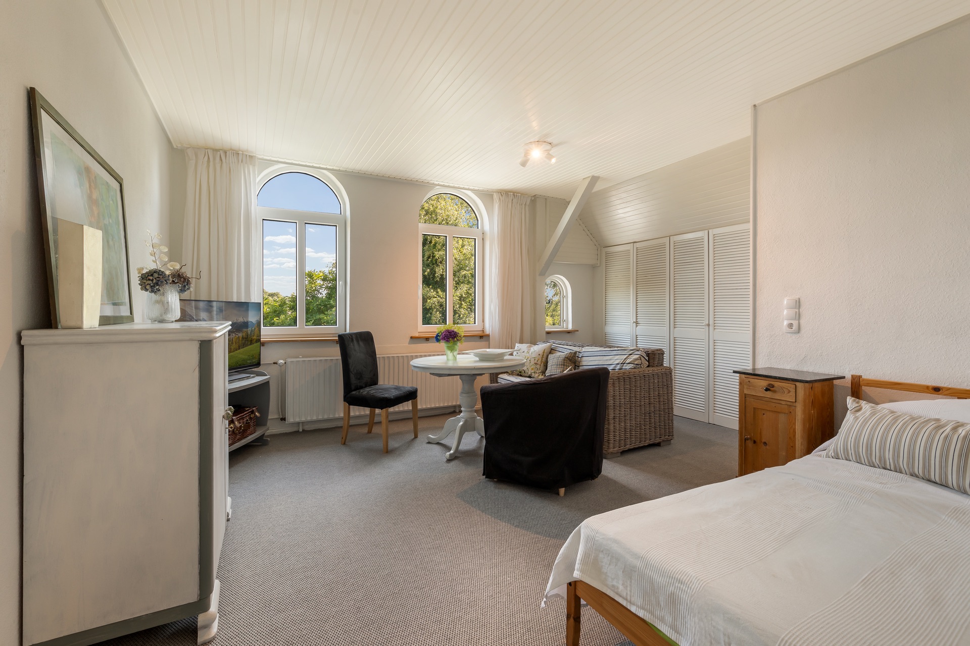 Helles Zimmer mit Bett, Sitzbereich, Esstisch und großen Fenstern mit Blick ins Grüne.
