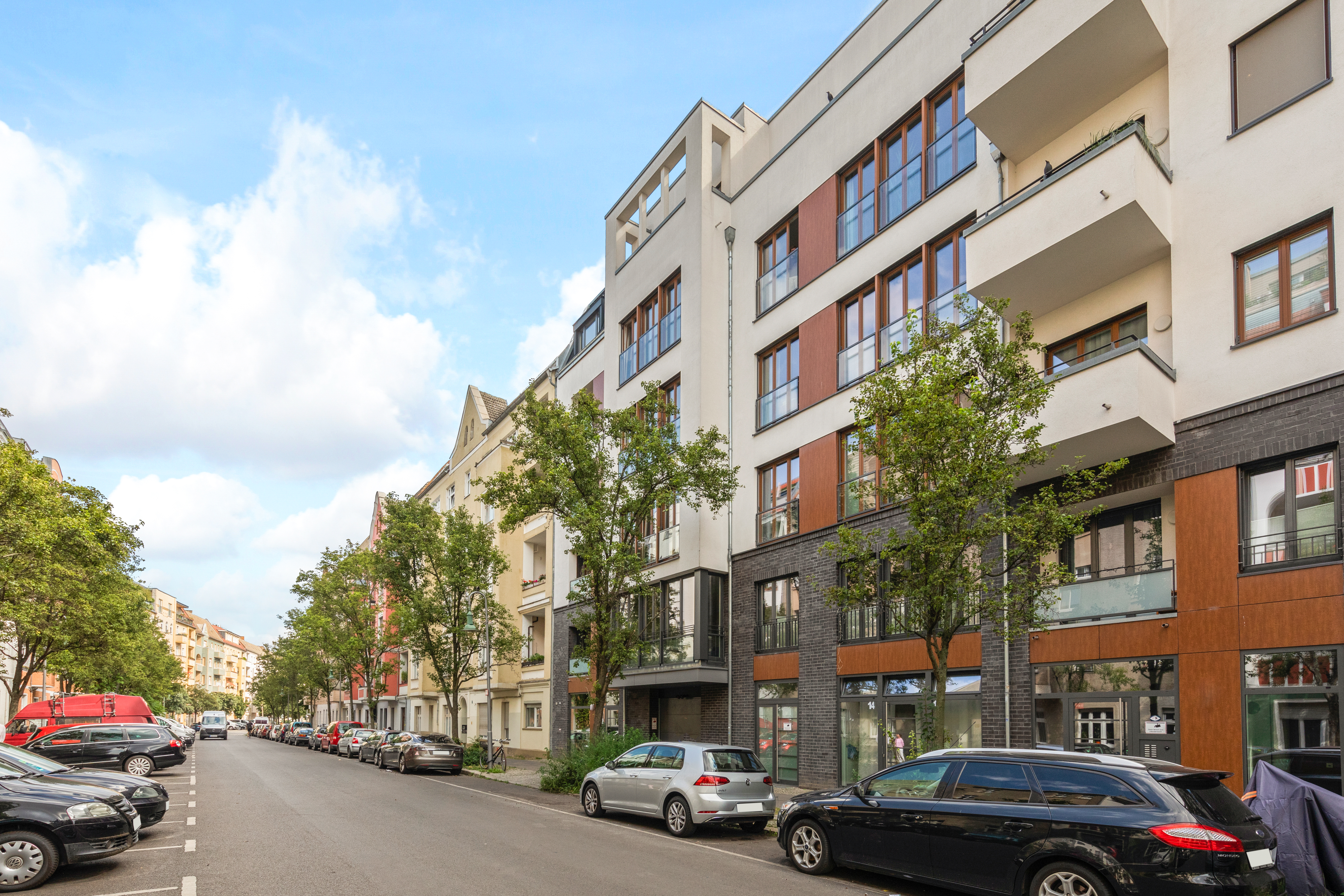 Moderne Wohngebäude mit Balkonen an einer Straße mit parkenden Autos und Bäumen.