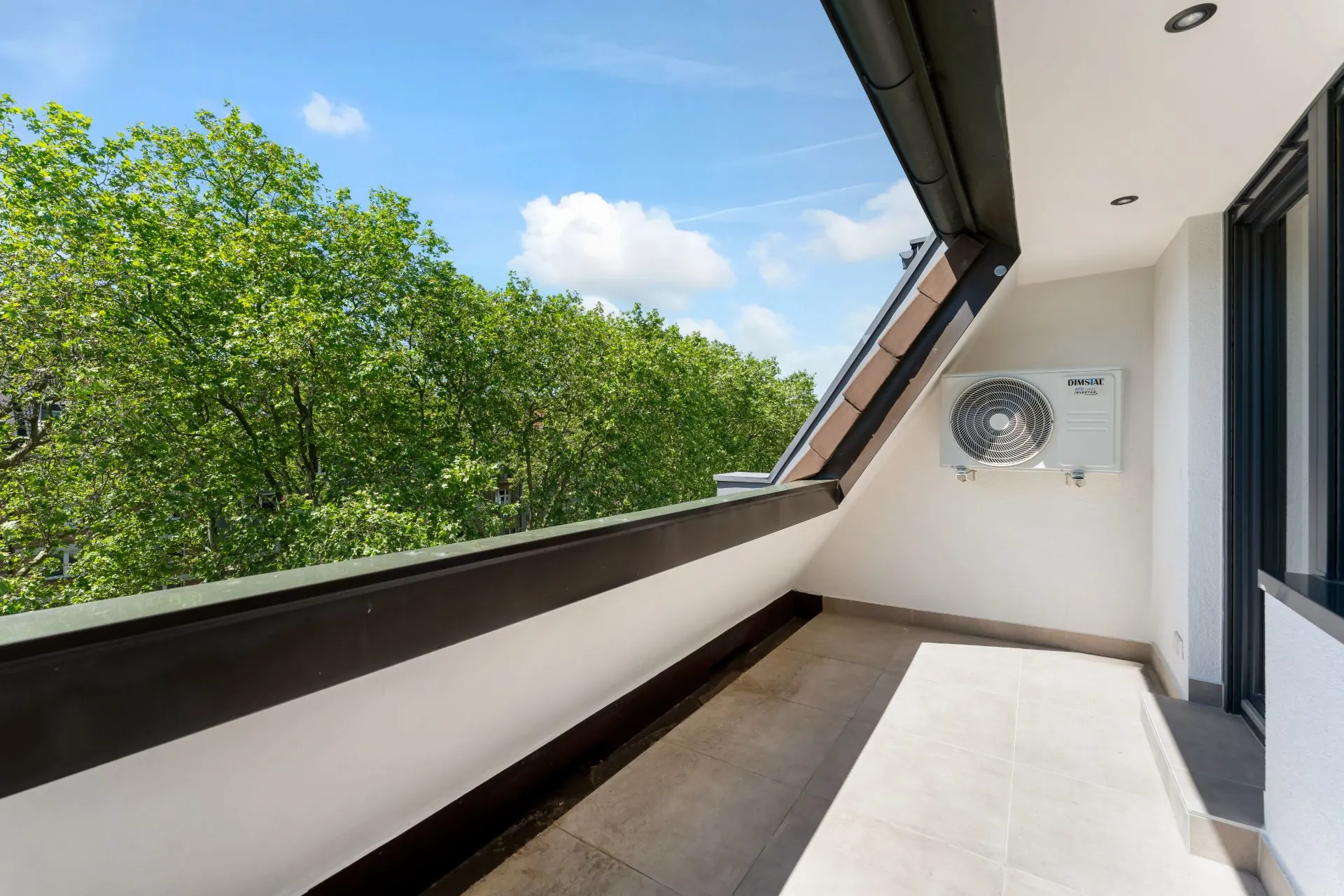 Balkon mit Fliesenboden, Wandklimaanlage und Blick auf grüne Bäume.
