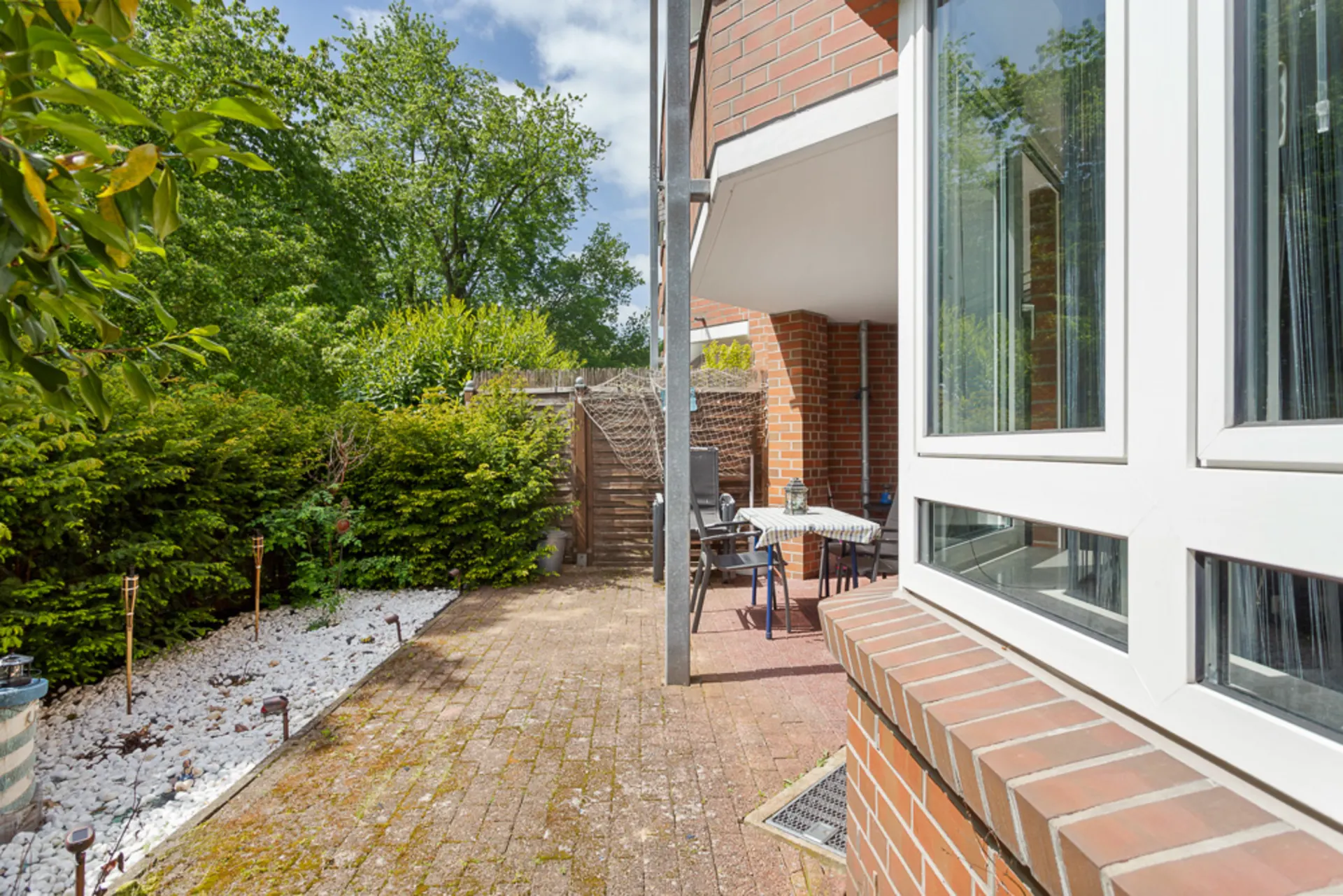 Gepflasterte Terrasse mit Tisch und Stühlen, umgeben von grünen Büschen und weißen Steinen.