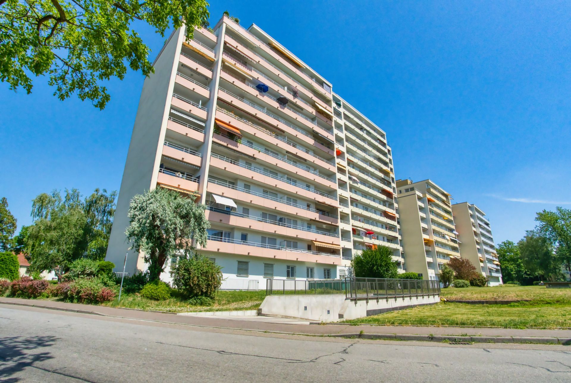 Mehrstöckiges Wohnhaus mit Balkonen und Grünflächen, an einem sonnigen Tag aufgenommen.