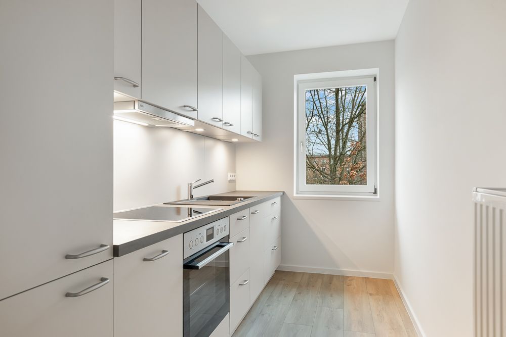 Moderne, schmale Einbauküche mit weißen Schränken und Fenster, hellem Boden und Elektroherd.