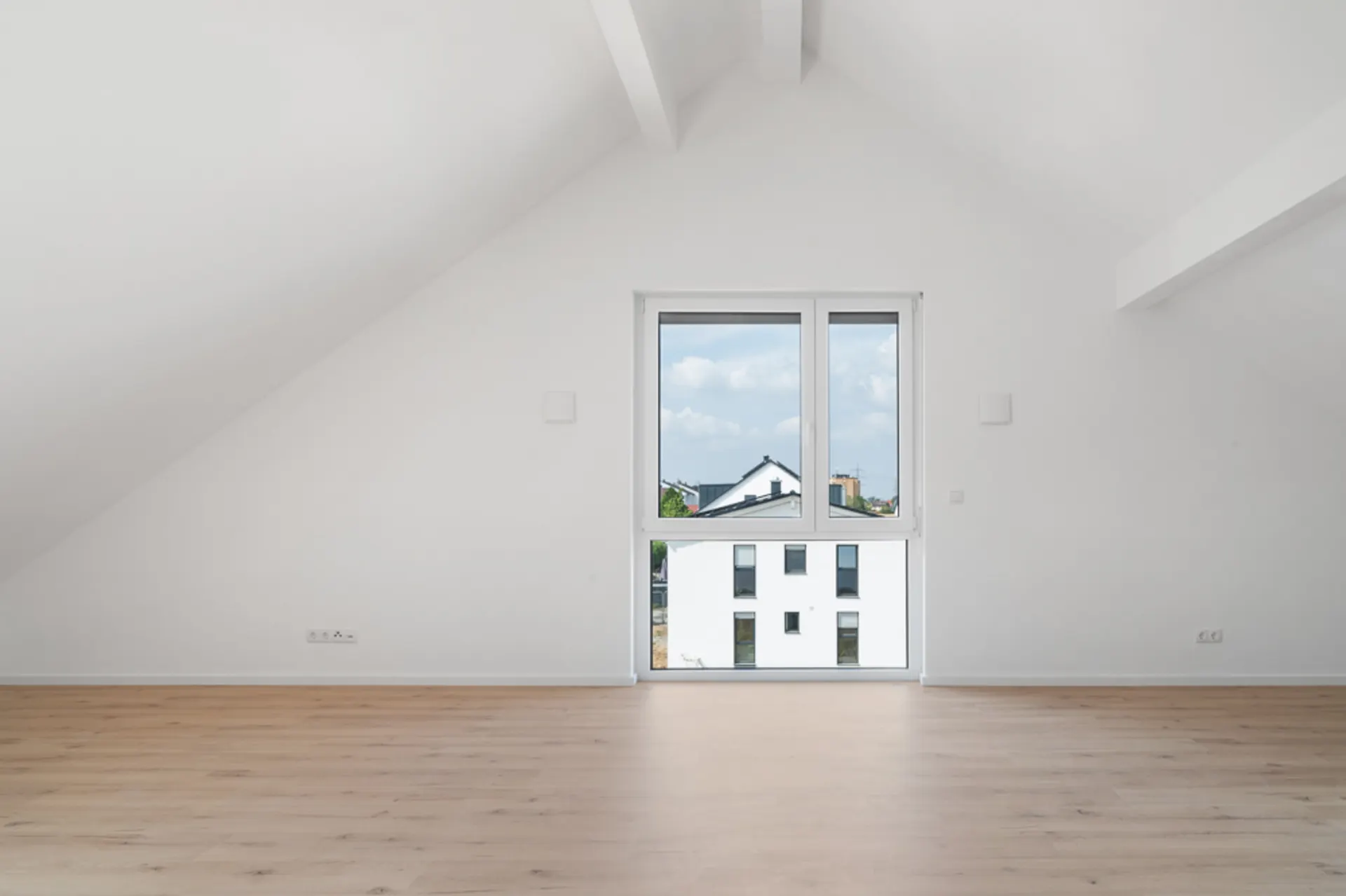 Heller, leerer Raum mit schräger Decke, großem Fenster und hellem Holzboden.