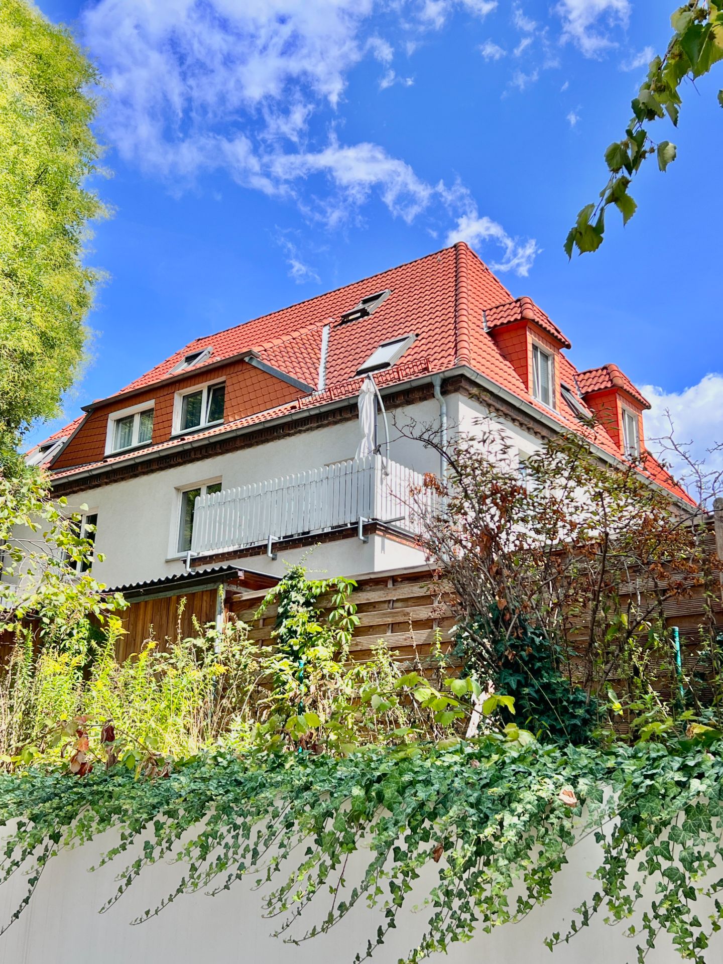 Modernes Wohnhaus mit rotem Dach, Balkon und viel Grün im Vordergrund.