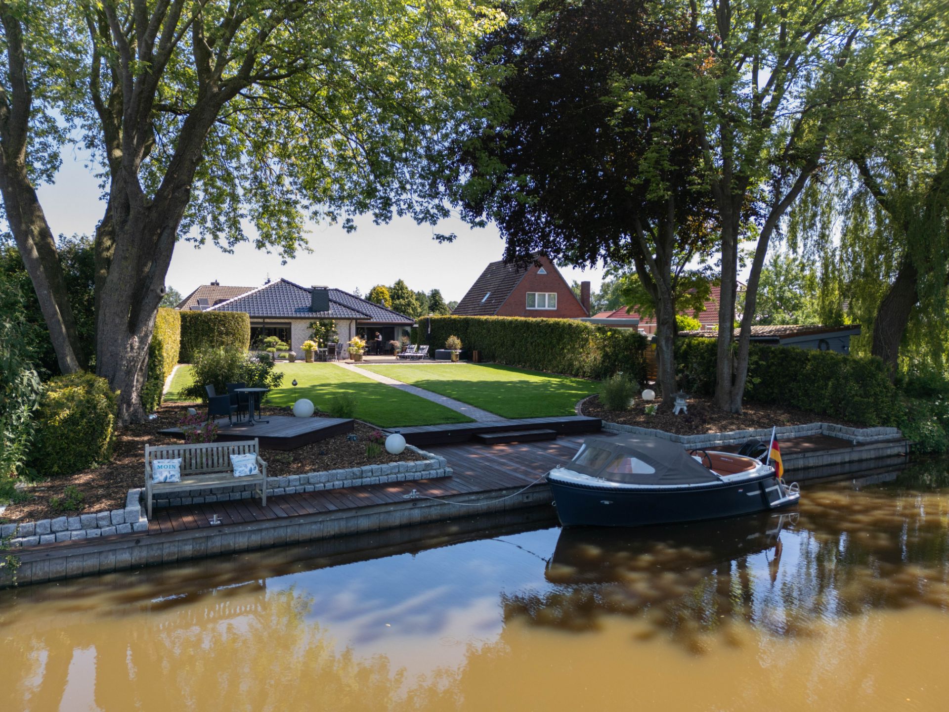 Garten mit Bootssteg, Sitzbank, Rasenfläche und Haus im Hintergrund an einem Fluss.