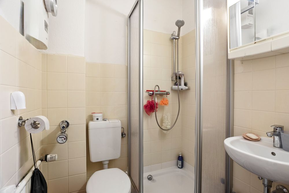Kleines Bad mit Toilette, Dusche und Waschbecken, beige Fliesen an den Wänden.