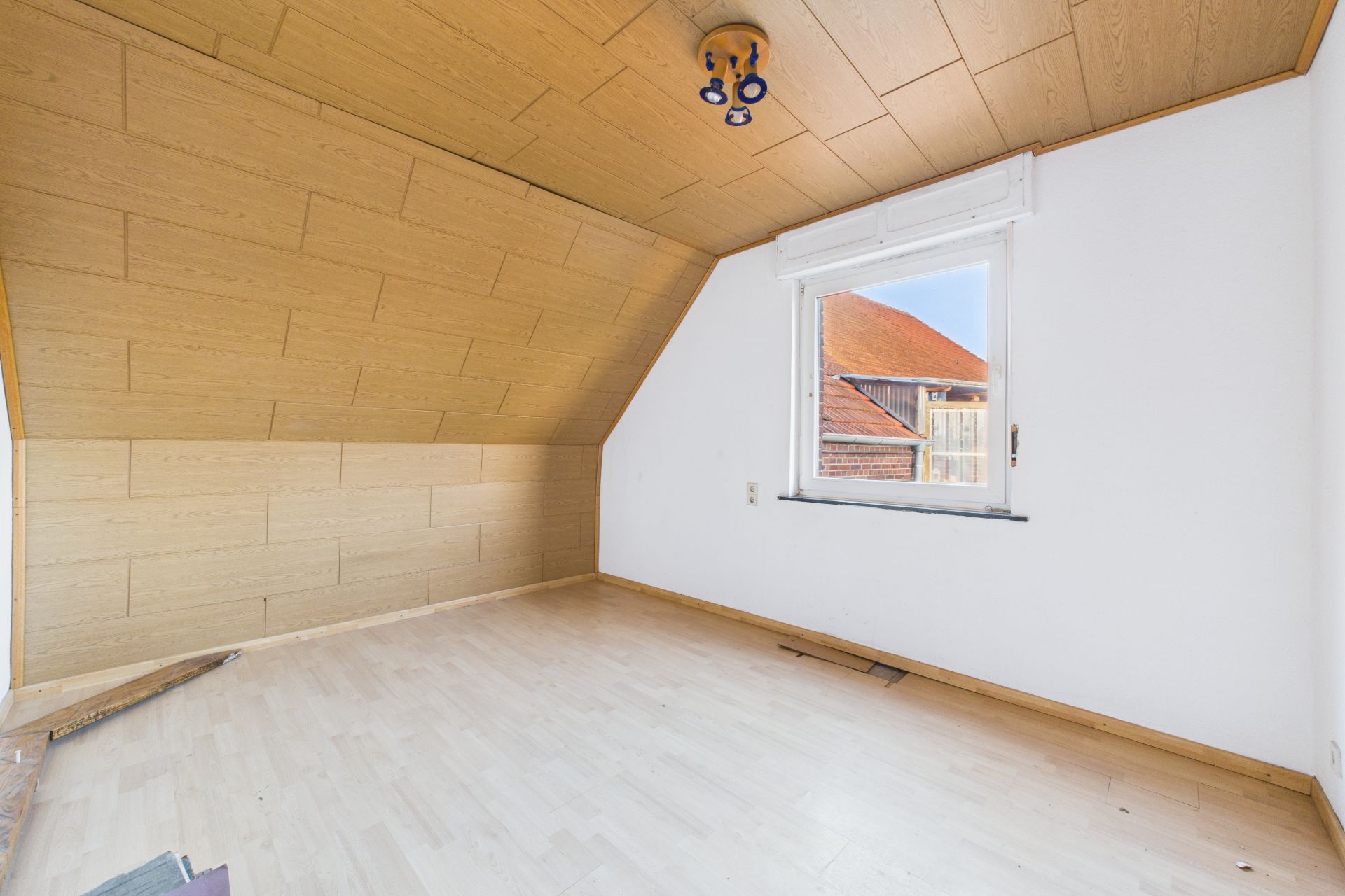 Leerer Raum mit schräger Holzdecke, weißer Wand und Fenster mit Blick auf ein Hausdach.