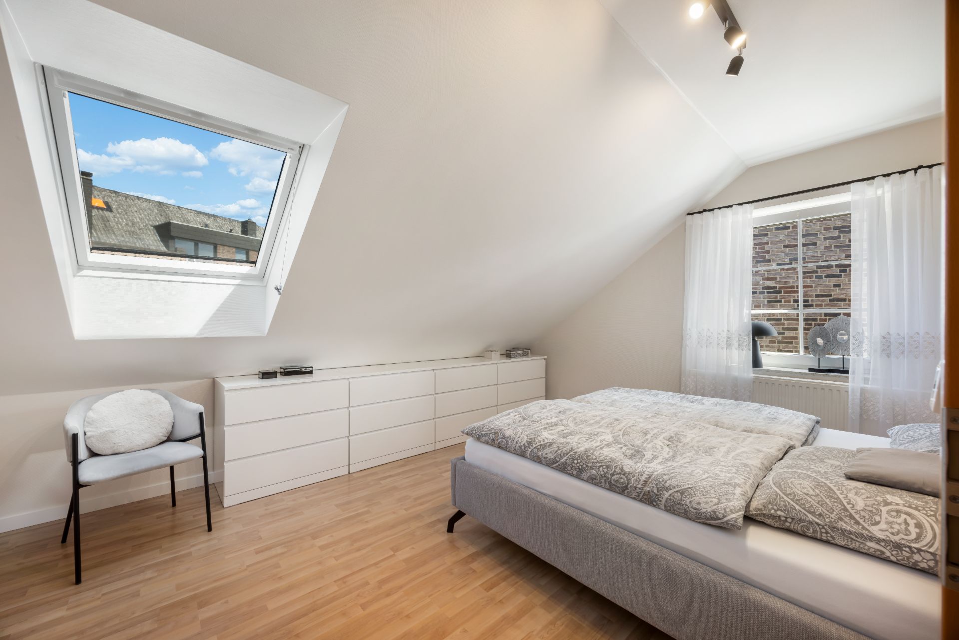Helles Schlafzimmer mit Bett, Kommode, Stuhl, Dachfenster und Fenster mit Vorhängen.