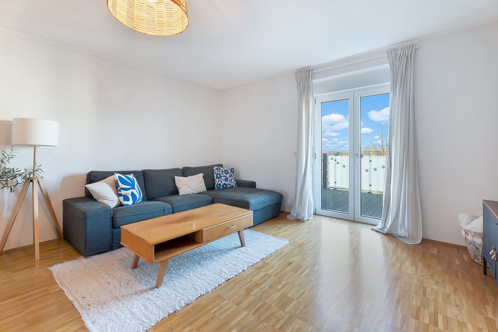 Helles Wohnzimmer mit grauem Sofa, Holztisch, Stehlampe und Zugang zu einem Balkon.