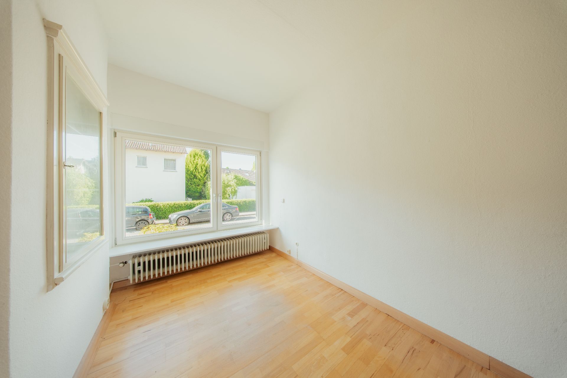 Heller leerer Raum mit Holzboden, großen Fenstern und Heizkörper unter dem Fenster.