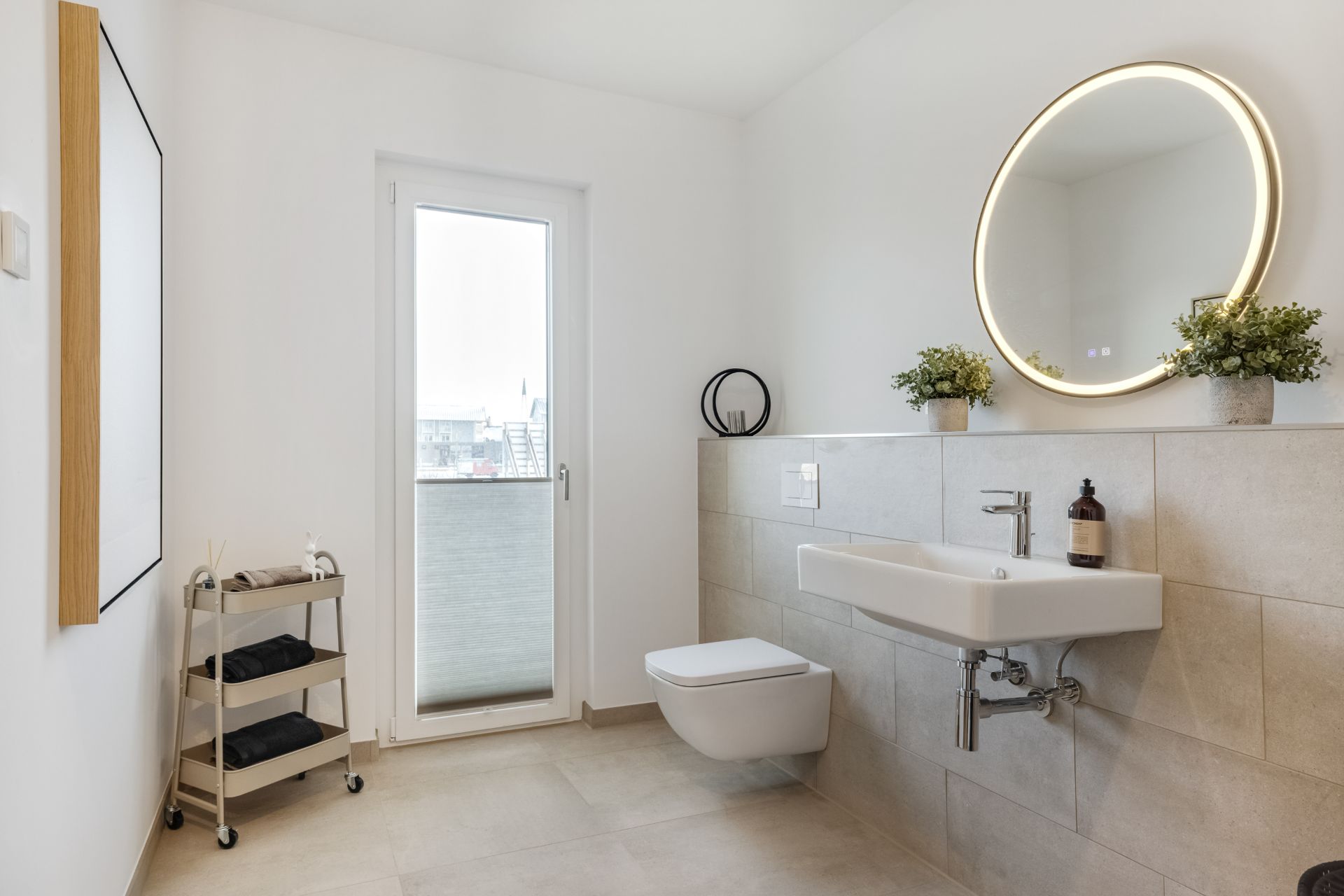 Modernes Badezimmer mit rundem Spiegel, Waschbecken, WC und Regalwagen neben einer Glastür.