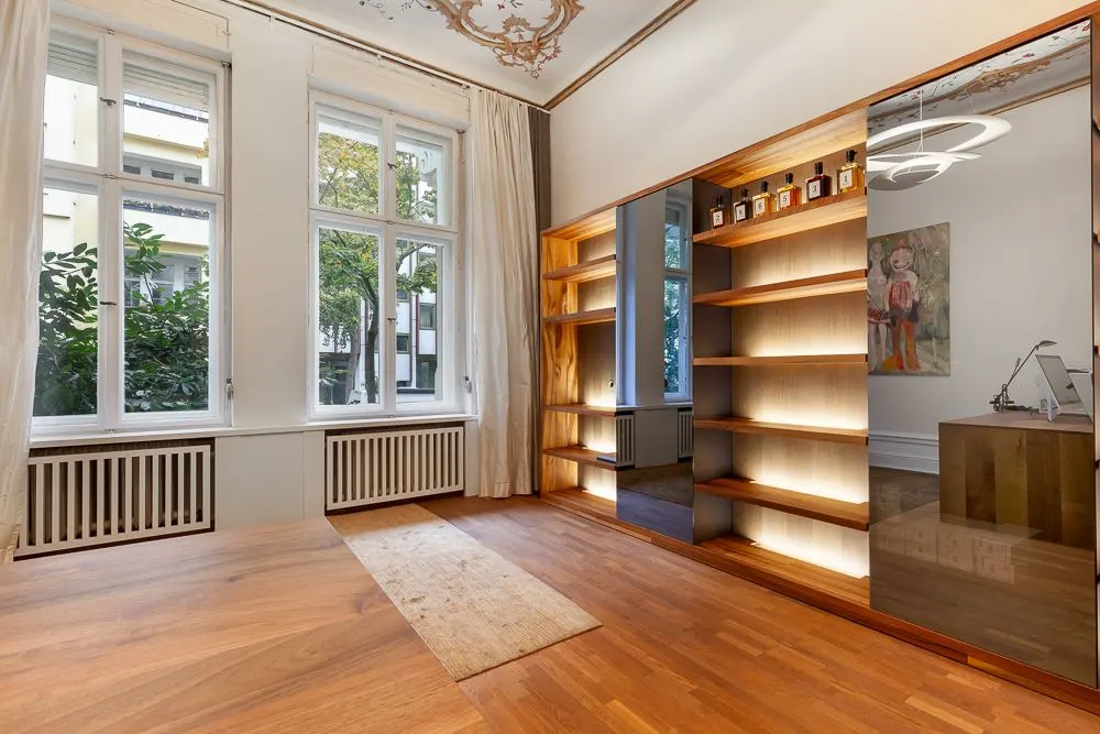 Heller Raum mit Holzboden, großen Fenstern, Regal mit Beleuchtung und Deckenverzierung.