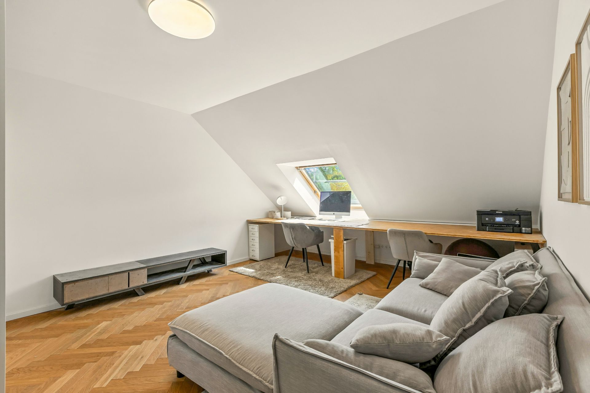 Helles Zimmer mit Sofa, Schreibtisch am Fenster, zwei Stühlen und Parkettboden.