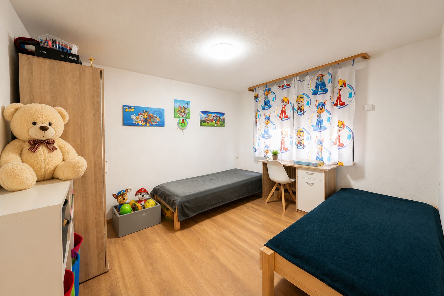 Kinderzimmer mit zwei Einzelbetten, Spielzeug, Schreibtisch und bunten Vorhängen.
