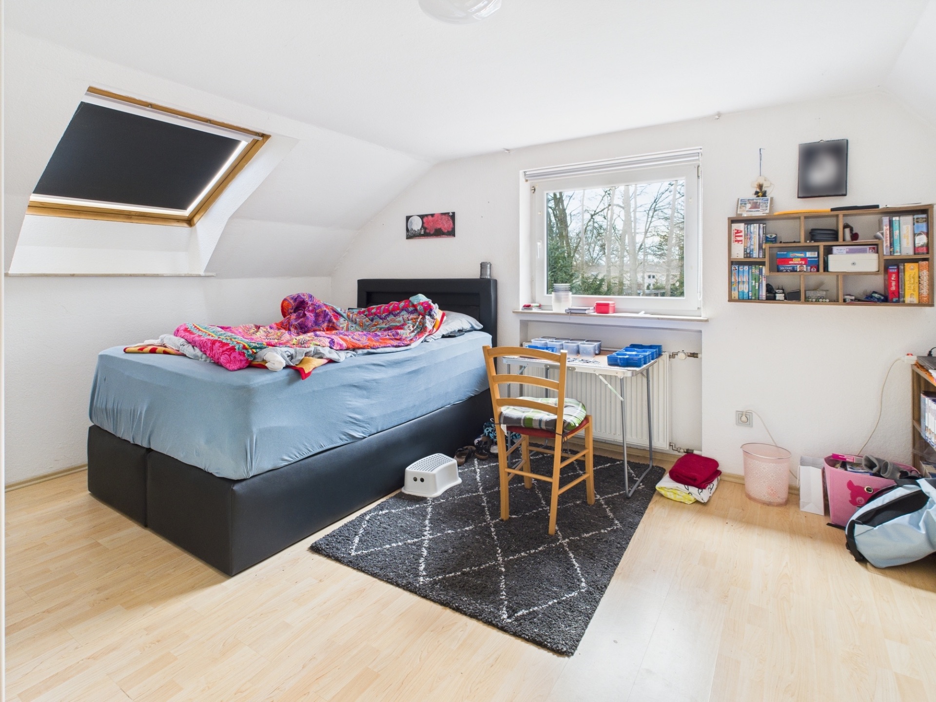 Helles Schlafzimmer mit Bett, Schreibtisch, Stuhl, Regal mit Spielen und Dachfenster.
