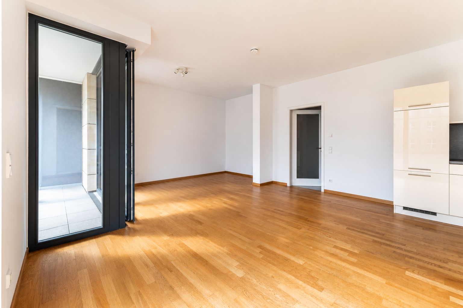 Helles, leeres Wohnzimmer mit Parkettboden und Zugang zu einem Balkon.