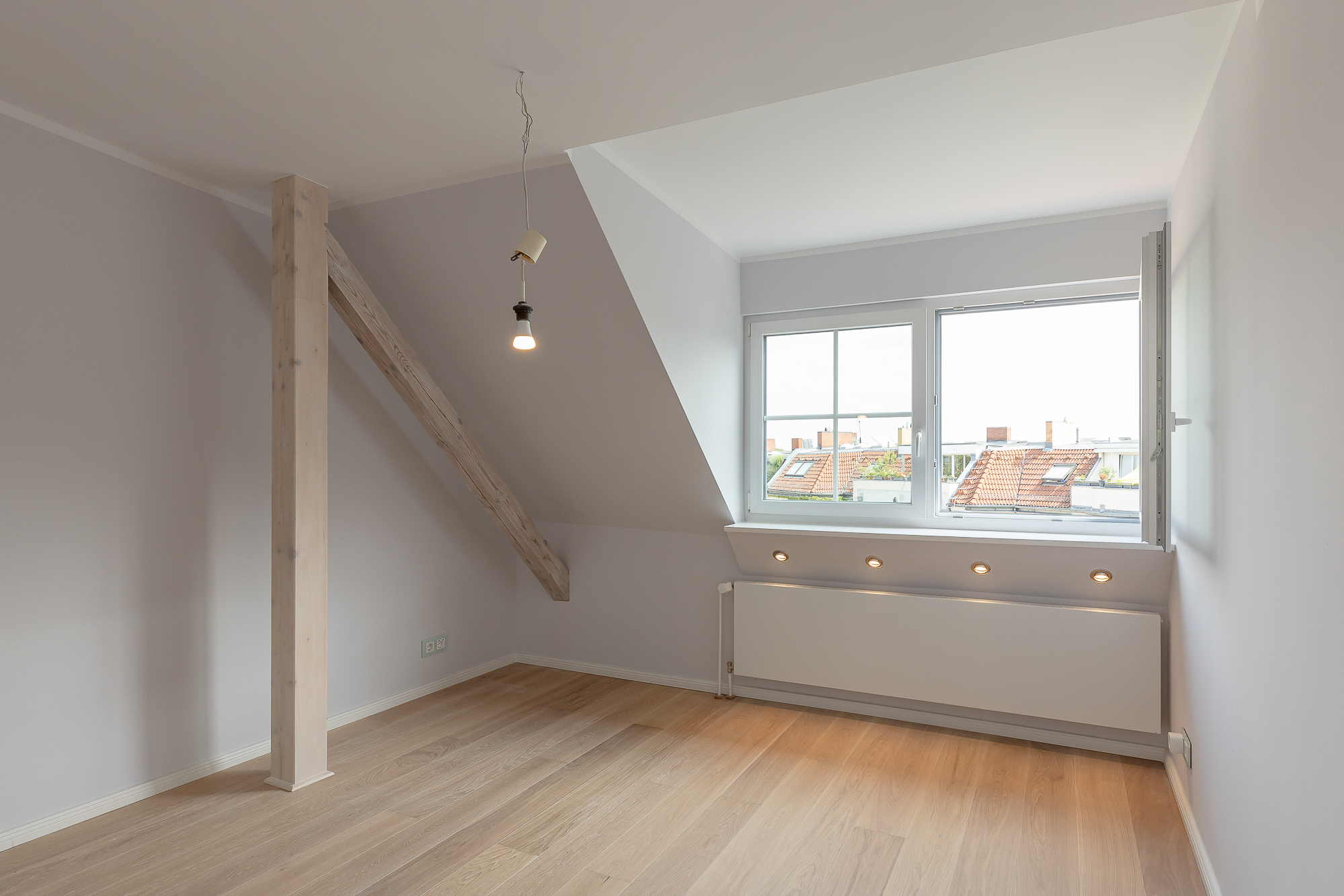 Helles Dachzimmer mit Holzfußboden, schräger Decke, Holzbalken und Fenster mit Blick nach draußen.