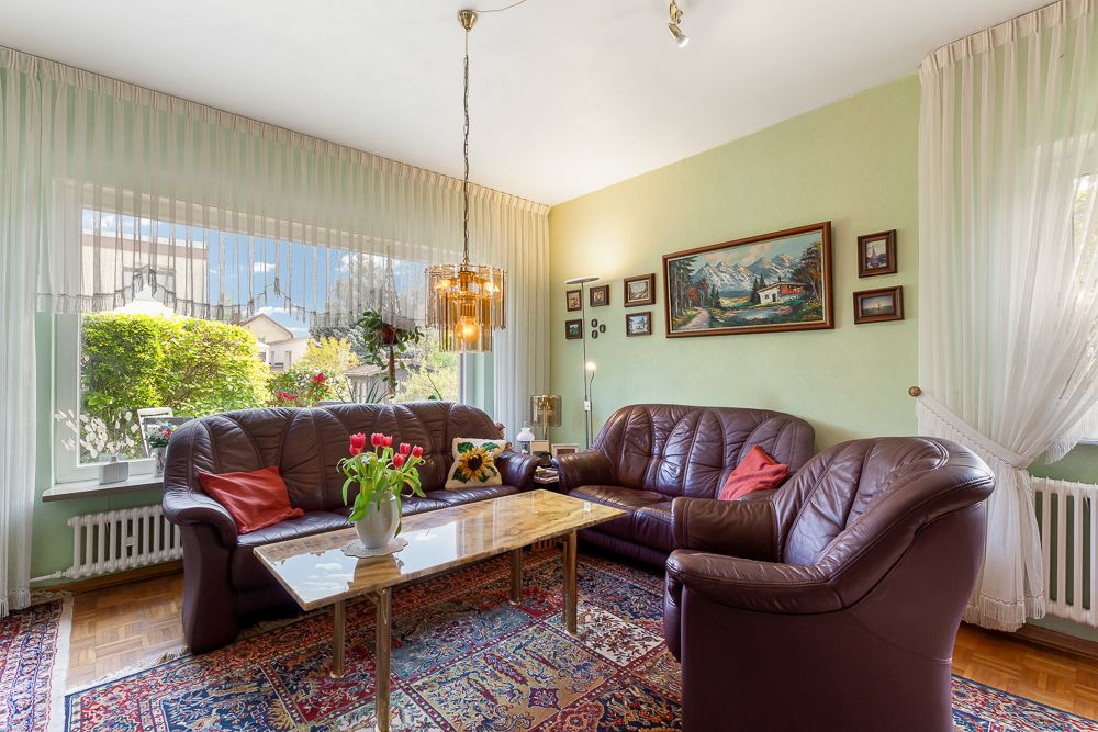Wohnzimmer mit braunen Ledersofas, Glastisch, buntem Teppich und großen Fenstern mit Gardinen.