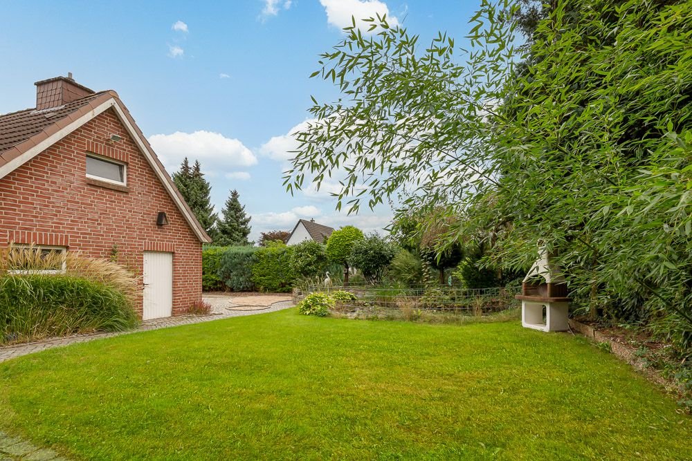 Gepflegter Garten mit Rasen, Grillplatz, Ziegelgebäude und vielen Bäumen.