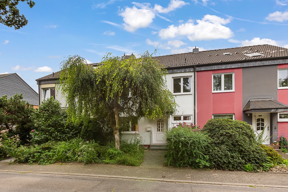 Reihenhaus mit weißer Fassade, Vorgarten, Bäumen und Sträuchern an einer ruhigen Straße.