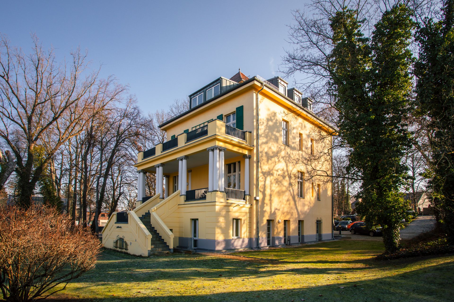 Gelbes Wohnhaus mit Säulen, Balkon und Treppe, umgeben von Bäumen.