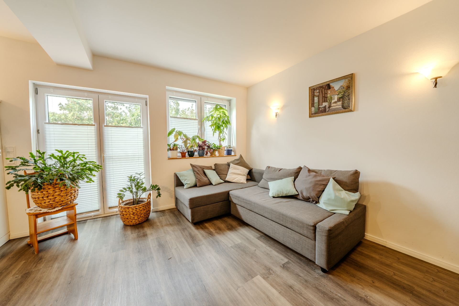 Helles Wohnzimmer mit Ecksofa, Pflanzen, Holzboden und großen Fenstern mit Jalousien.