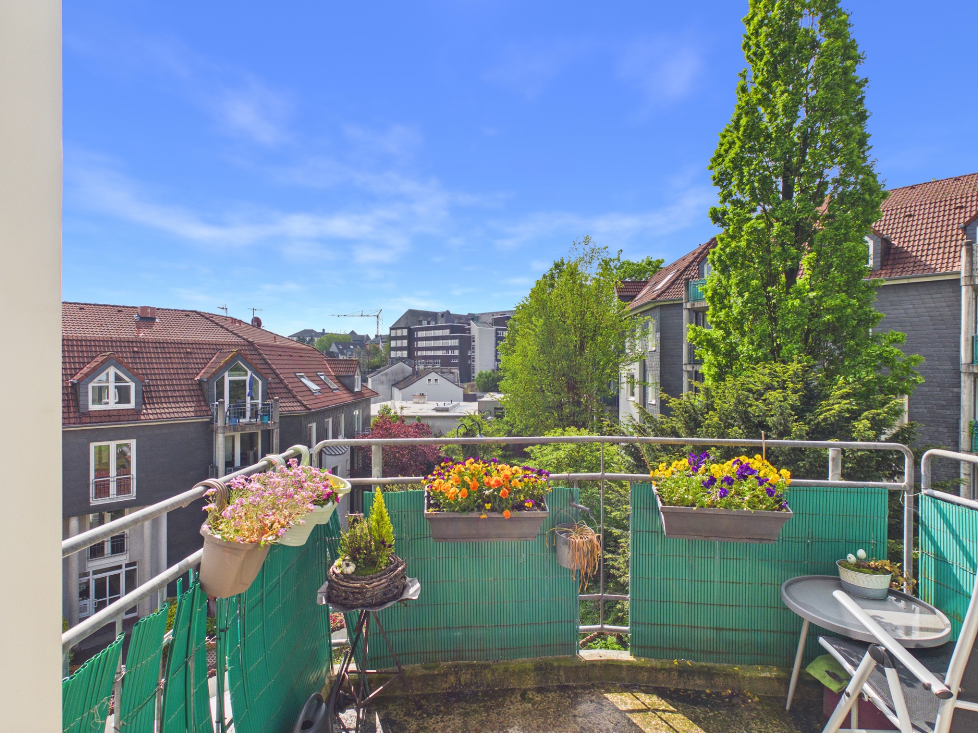 Balkon mit Blumenkästen, kleinem Tisch und Blick auf Häuser und Bäume.