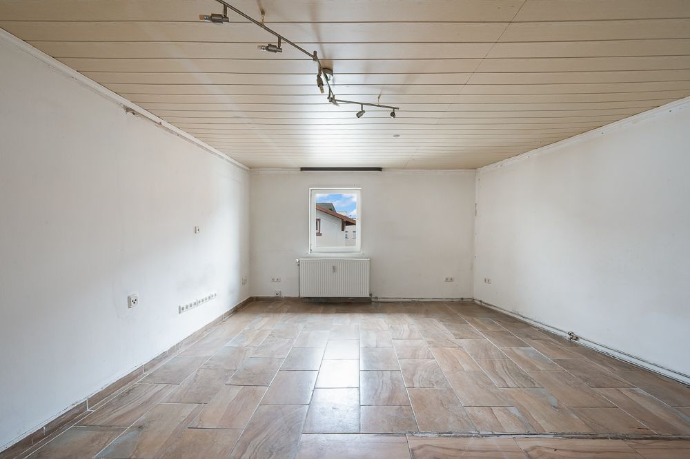 Leerer Raum mit Holzboden, weißem Wänden, Deckenleuchte und Fenster mit Heizkörper darunter.