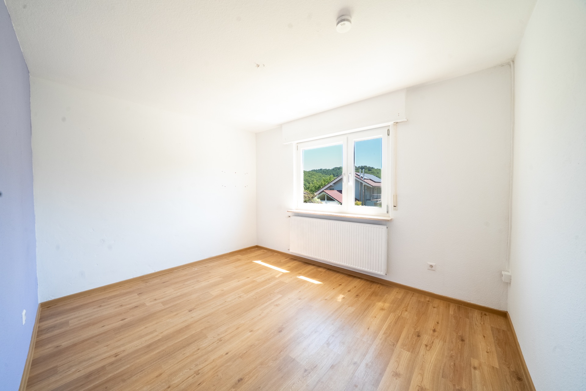 Helles, leeres Zimmer mit Holzfußboden und großem Fenster mit Blick ins Grüne.