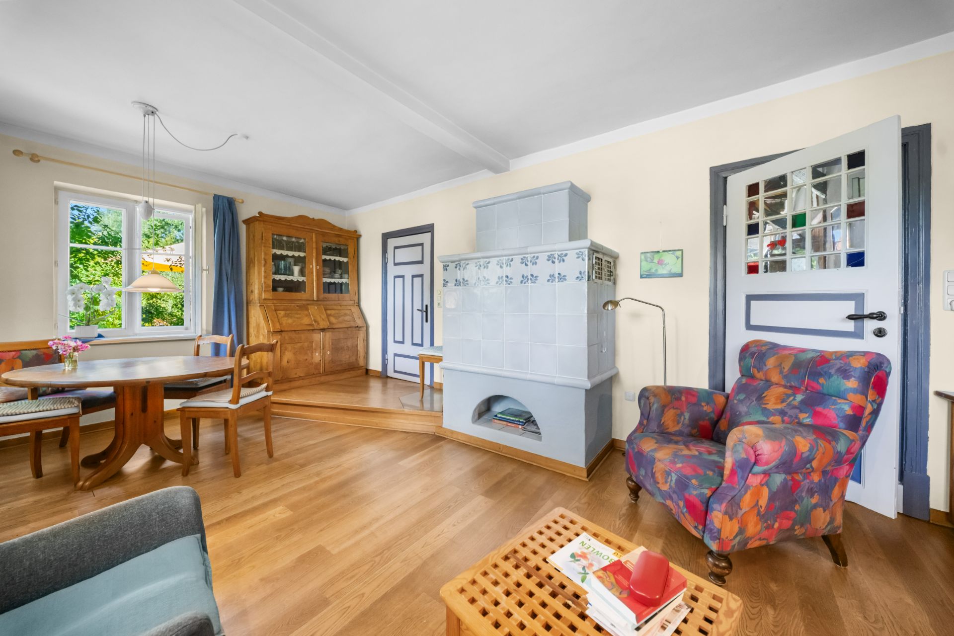 Wohnzimmer mit Kaminofen, buntem Sessel, Holztisch und großem Fenster mit Blick ins Grüne.