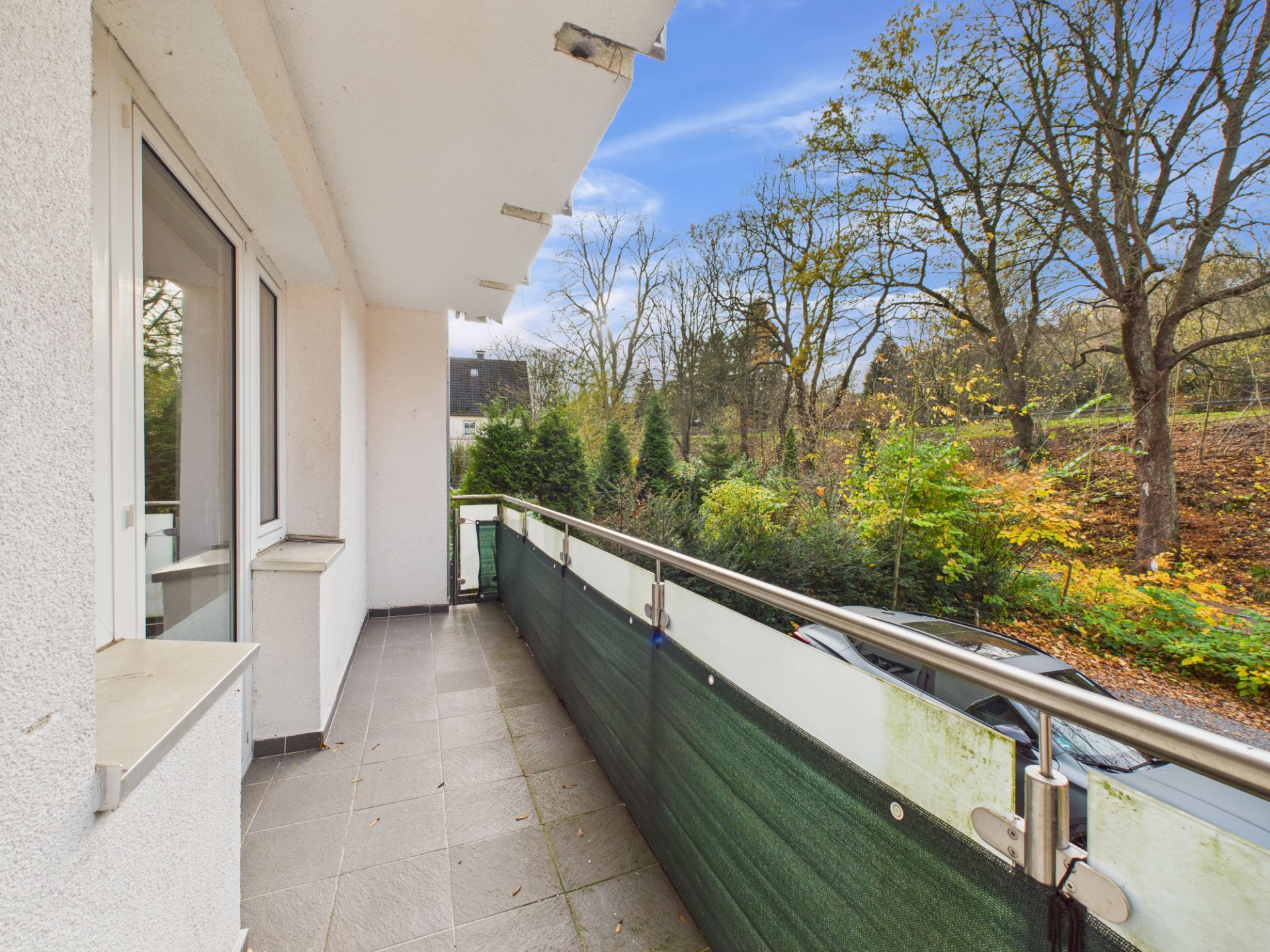 Balkon mit Fliesenboden und grünem Sichtschutz, Blick auf Bäume und Sträucher.
