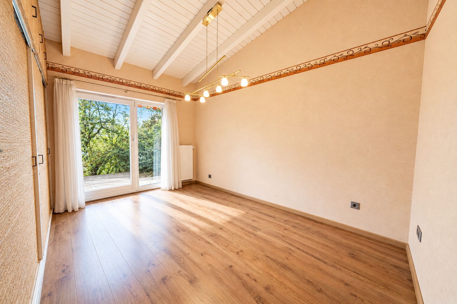 Leerer Raum mit Holzfußboden, Deckenbalken und großem Fenster zur Terrasse.