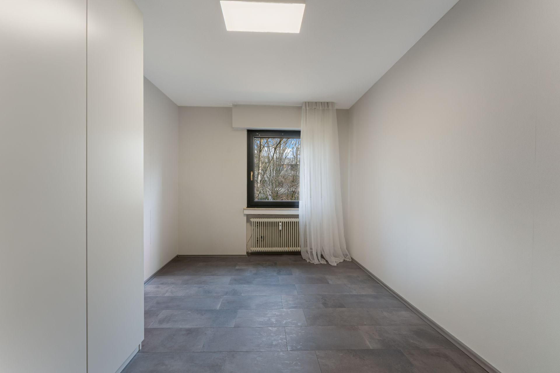 Leerer, heller Raum mit Fliesenboden, Fenster, Heizung und weißem Vorhang.