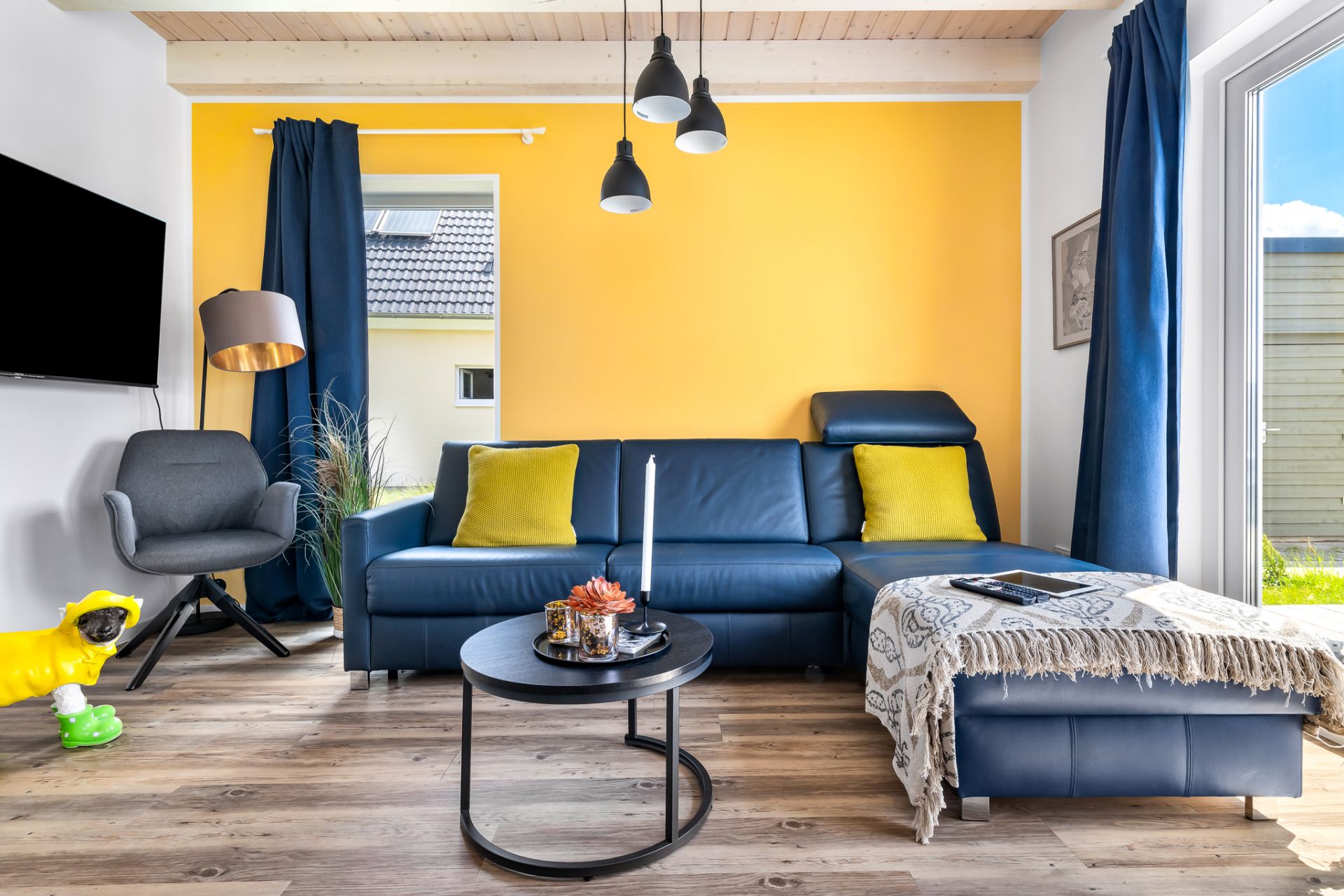 Wohnzimmer mit blauer Couch, gelben Kissen, gelber Wand und Couchtisch, viel Tageslicht.